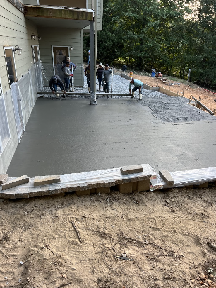 Crew of six pouring and screeding a large backyard concrete patio, with stone block retaining wall forms and a wooded backdrop