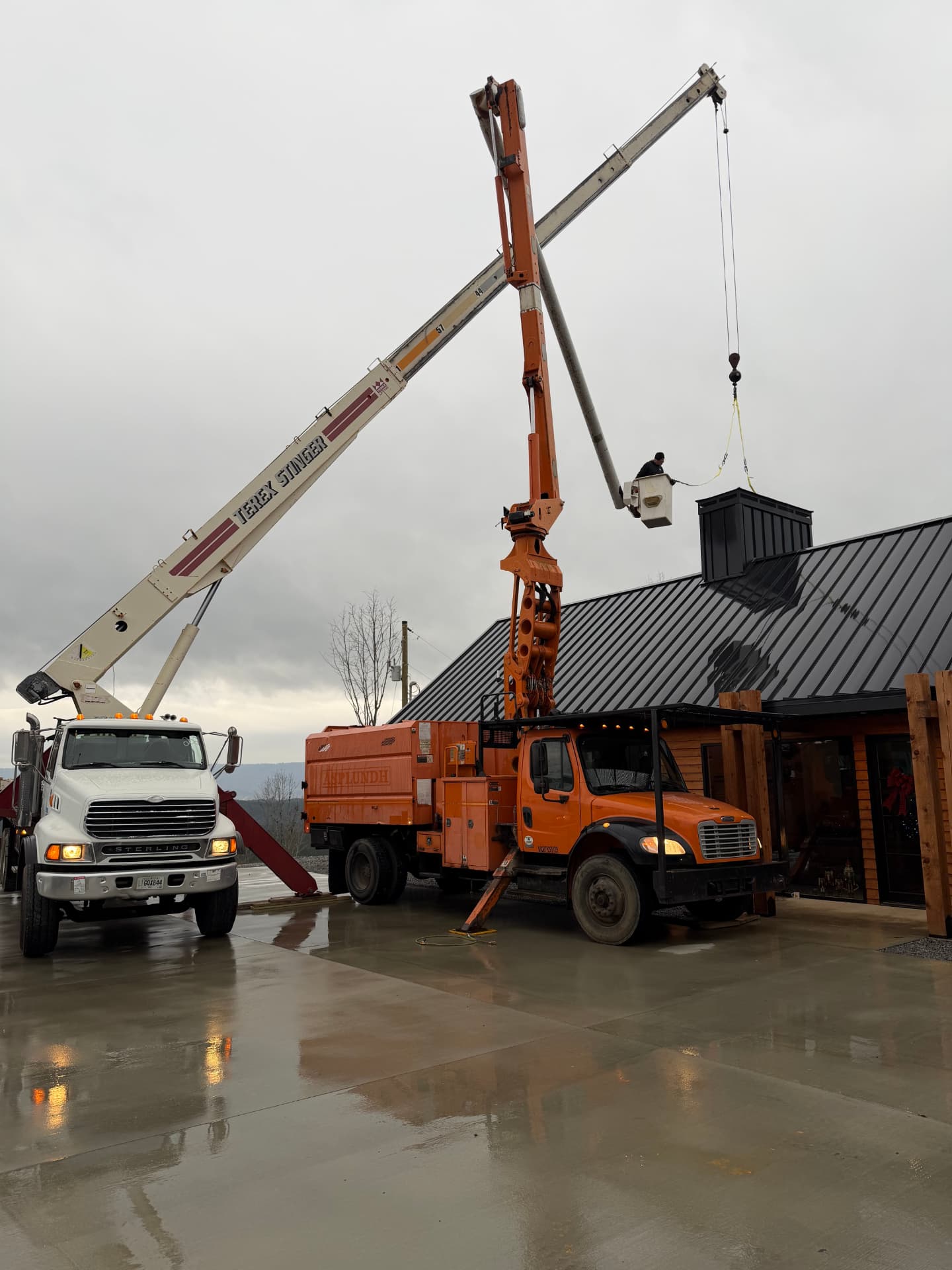 Crane truck and bucket truck lifting material beside a building on a wet day