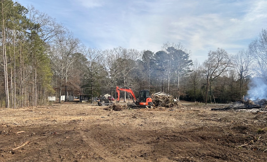 Orange Kubota excavator clearing a rural lot, with a burn pile and cleared logs behind it