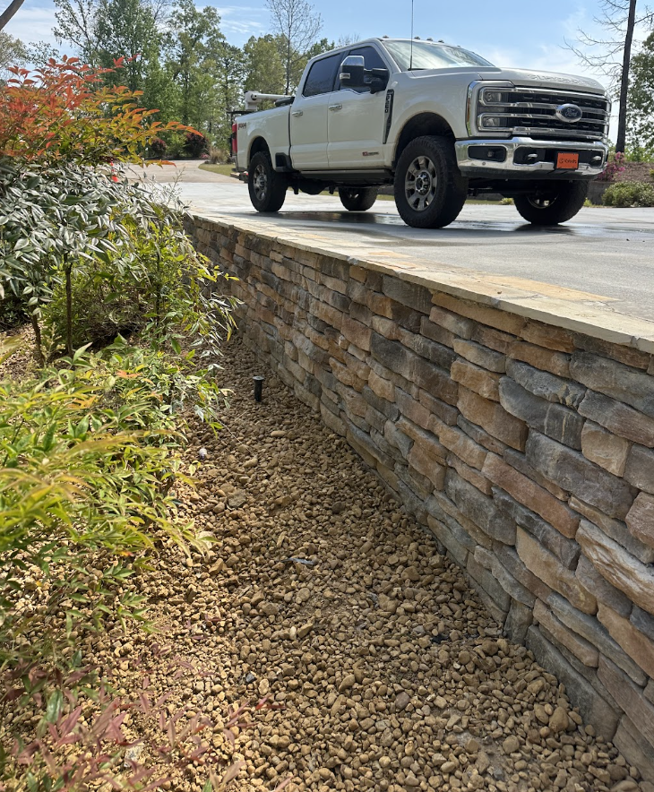 Stacked stone retaining wall with a flat stone cap, lush landscaping and gravel bed, with a white Ford truck parked on the driveway above